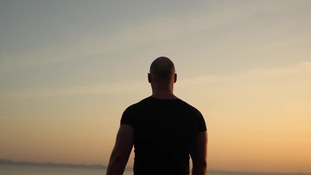 closeup picture from back of muscular strong bald man in black T-shirt standing on top of hill looking at sea putting arms up in the air enjoying pastime on nature slow motion