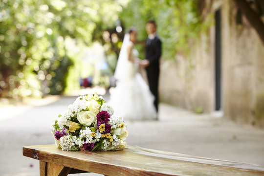 bouquet on wooden bench with young bride and groom in the background. 