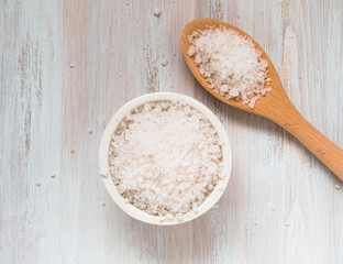 White bowl with large sea salt and wooden spoon on white wooden table. Top view.