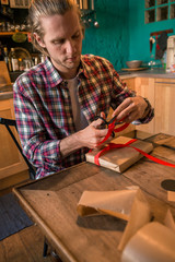 Young adult man with long blonde hair wrapping christmas present with red ribbon seated on wooden table in cozy apartment indoor.