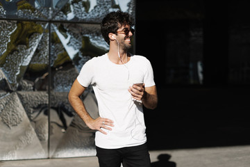 Fototapeta premium Young caucasian man in a blank white t-shirt enjoys the city walk with the music he listens on a mobile phone. Handsome bearded guy wearing sunglasses stands with a smartphone in his hands.
