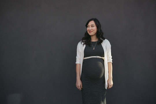 Pretty young pregnant woman standing near grey wall outside smiling