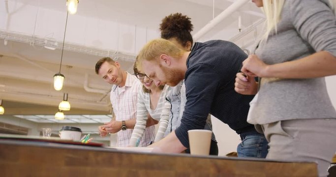 Group of people in casual clothes standing near table in office, looking at draft and smiling after discussion of new project.