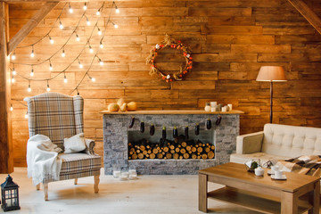 Home comfort. Armchair near the fireplace with firewood. Photo of interior of room with a wooden wall, wreath and garlands, Christmas atmosphere - obrazy, fototapety, plakaty