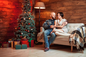 Obraz premium Beautiful couple sitting on the couch embrace in a decorated festive interior with a Christmas tree. The concept of a family celebration, new year's eve