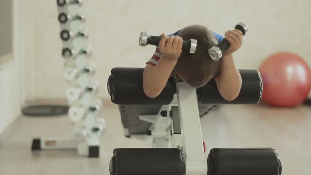 Young boy doing heavy dumbbell exercise for biceps. Baby working out with dumbbells on training apparatus. Strength and motivation. The concept of a healthy lifestyle. GYM
