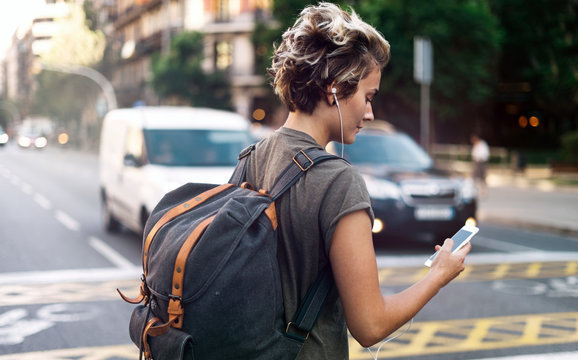 Student girl is usinf smartphone to find the address of the hostel. Young tomboy girl is listening to the music on her mobile phone while walking the street on a blurred urban background.