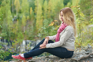 Young woman with long blonde hair having a rest on the edge of the cliff against picturesque forest.