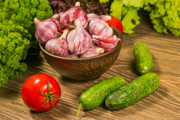 A full bowl with garlic bulbs. Tomatoes and cucumbers on a wooden table. Green salad in the background.