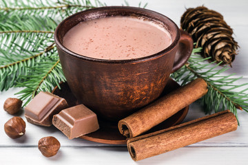 Christmas cup of hot cocoa with milk, broken chocolate cubes, hazelnut and cinnamon sticks, christmas tree branches, pine cone on white wooden planks