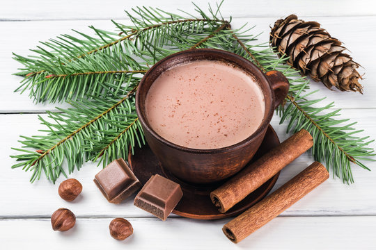 Christmas cup of hot cocoa with milk, broken chocolate cubes, hazelnut and cinnamon sticks, christmas tree branches, pine cone on white wooden planks