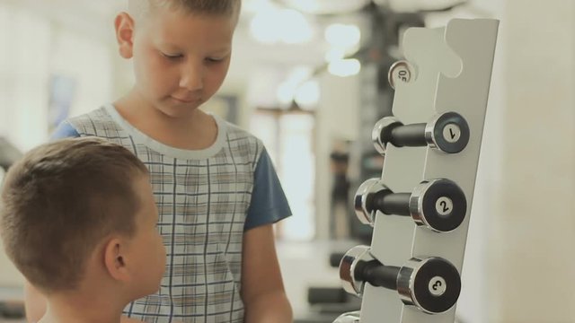 The older boy helps the younger to choose dumbbells for his workout in the gym. The concept of sporting motivation to increase. Teenager cares about baby's body and health from the childhood