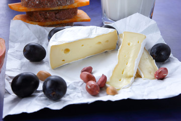 Camembert cheese, orange marble cheese, a glass of milk, pomegranate, olives, peanut grains, dark blue grapes and bread on white fabric and dark blue background, cheese background in retro style
