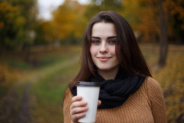 Young woman wearing knitted sweater walking in the autumn park and drinking take away coffee in paper cup. Breakfast on the go, warming concept