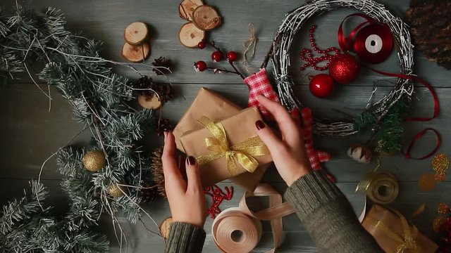 A top-down plan. Fully visible the table with the decorations. Female hands put and finalize Christmas gift wrapped in craftool paper on a wooden table. Bandaging tape and tied in a bow.