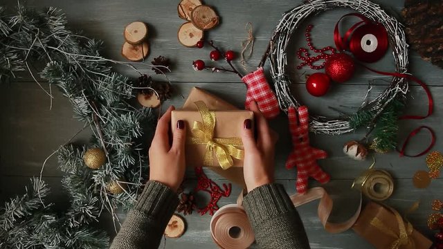 A top-down plan. Fully visible the table with the decorations. Female hands put and finalize Christmas gift wrapped in craftool paper on a wooden table. Bandaging tape and tied in a bow.