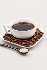 White coffee cup with coffee, plate with coffee beans and black teaspoon with brown sugar
