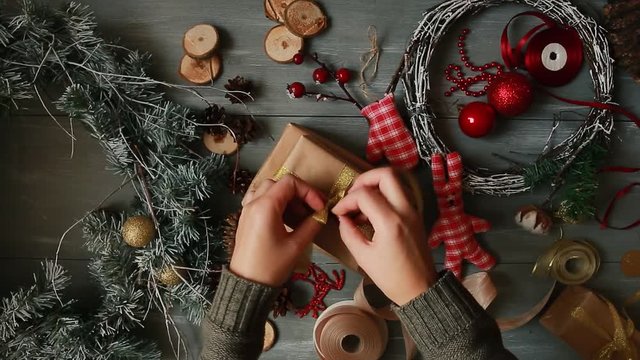A top-down plan. Fully visible the table with the decorations. Female hands put and finalize Christmas gift wrapped in craftool paper on a wooden table. Bandaging tape and tied in a bow.