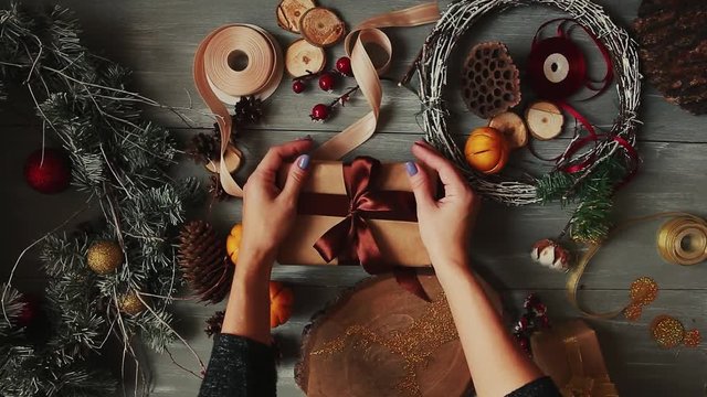 A top-down plan. Fully visible the table with the decorations. Female hands put and finalize Christmas gift wrapped in craftool paper on a wooden table. Bandaging tape and tied in a bow.