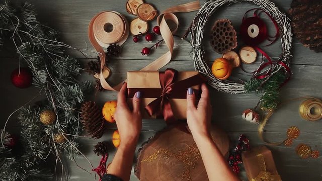 A top-down plan. Fully visible the table with the decorations. Female hands put and finalize Christmas gift wrapped in craftool paper on a wooden table. Bandaging tape and tied in a bow.
