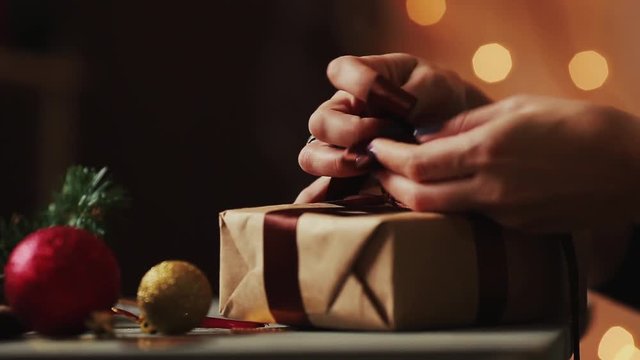 Close-up on the background of Christmas lights women's hands tying the ribbon on the gift of Kraft paper. The background is blurred in bokeh.