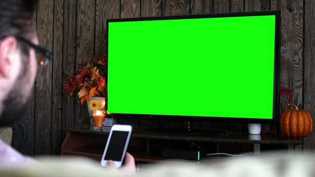Millennial male on smartphone while watching generic green screen TV in living room