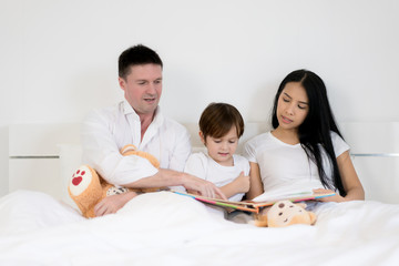 Asian child boy reading book together on bed in bedroom with father and mother. Happy family concept.