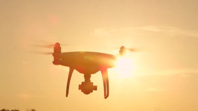 CLOSE UP LENS FLARE SILHOUETTE Small filming drone flying over golden sunset sky. White quadcopter drone with attached camera filming at golden light. Quadrocopter helicopter flying across sunrise sky