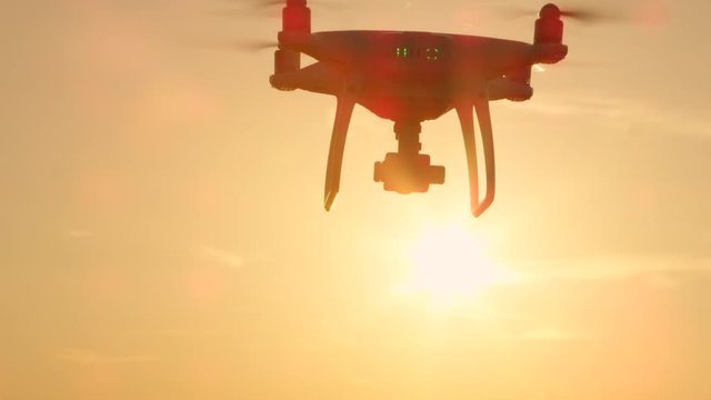 CLOSE UP LENS FLARE SILHOUETTE Small filming drone flying over golden sunset sky. White quadcopter drone with attached camera filming at golden light. Quadrocopter helicopter flying across sunrise sky