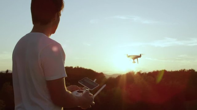 SLOW MOTION CLOSE UP LENS FLARE: Young man flying quadro drone with modern white RC transmitter. Male filmer controlling drone over RC radio controller. Man flying and filming with drone camera