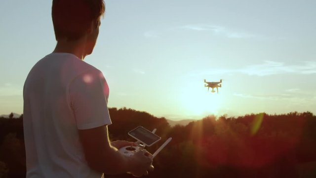 SLOW MOTION CLOSE UP LENS FLARE: Young man flying quadro drone with modern white RC transmitter. Male filmer controlling drone over RC radio controller. Man flying and filming with drone camera