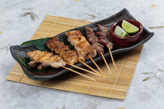 Yakitori (Japanese-Style Grilled Chicken Skewers) with chicken and internal organ served with sliced lime on black stone plate on makisu (Meal mat).