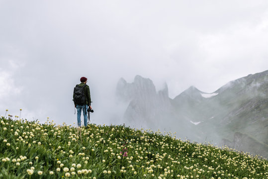 Male photographer wearing flannel shirt, olive jacket and jeans gazing at a distant mountain on an overcast day while standing in a field of flowers in Switzerland