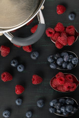 berry raspberries and blueberries, lies in four spoons on a black background, in the corner there is a cooking vessel