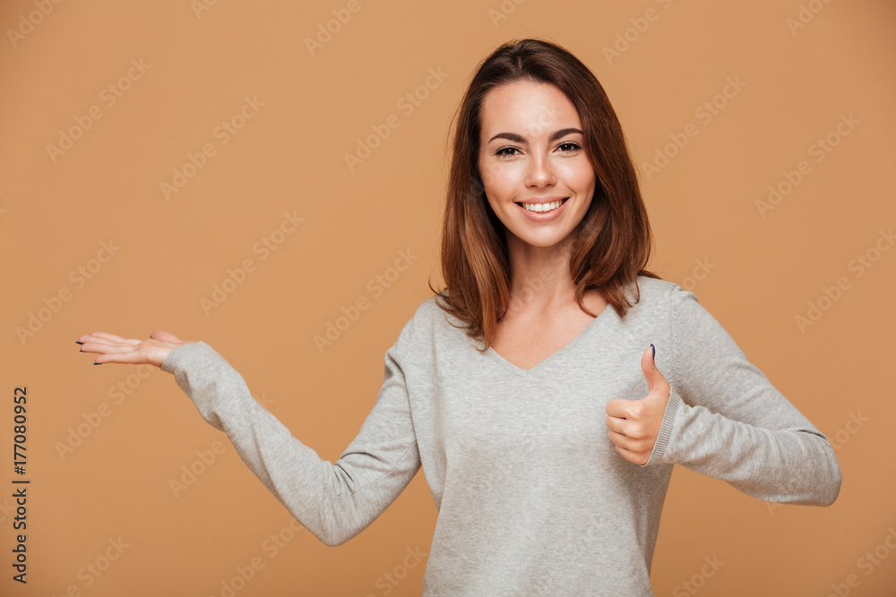 Poster Cheerful young brunette woman standing with empty palm, showing thumb up gesture