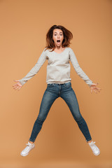 Full length photo of shocked young woman in gray blouse and jeans jumping over beige background