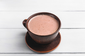 Hot cocoa with milk in brown clay cup, on table of white wooden planks