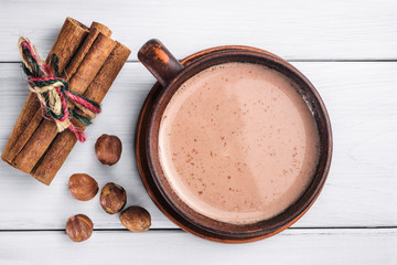 Hot cocoa with milk in brown clay cup, hazelnut and cinnamon sticks on table of white wooden planks, top view