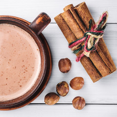Hot cocoa with milk in brown clay cup, hazelnut and cinnamon sticks on table of white wooden planks, top view