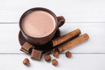 Photo sur Plexiglas Chocolat Hot cocoa with milk in brown clay cup, broken chocolate cubes, hazelnut and cinnamon sticks on white wooden planks