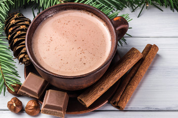 Christmas cup of hot cocoa with milk, broken chocolate cubes, hazelnut and cinnamon sticks, christmas tree branches, pine cone on white wooden planks