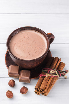 Hot cocoa with milk in brown clay cup, broken chocolate cubes, hazelnut and cinnamon sticks on white wooden planks