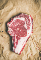 Flat-lay of raw uncooked prime beef meat dry-aged steak rib-eye on bone with seasoning on craft paper background, top view, copy space. Meat high-protein dinner concept