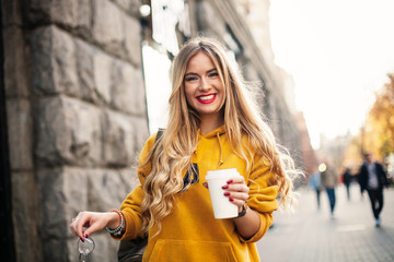 young stylish girl student wearing boyfrend jeans bright yellow sweetshot.She holds coffee to go. portrait of smiling girl in sunglasses and with bag posing in the street