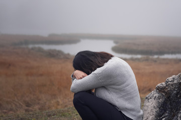 Depressive Frau lehnt sitzt am Fels Stein mit vor dem Gesicht verschränkten Armen düstere Stimmung