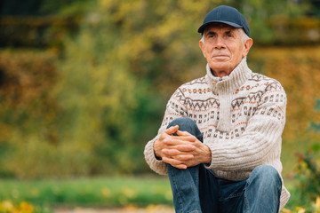 Portrait of pretty senior man in cap sitting in colorful autumn park. Pensioner resting outdoors. Beutiful fall.