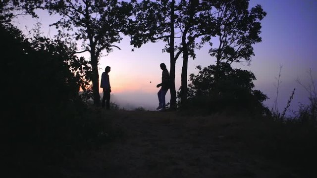 silhouettes of two tall slim guys playing footbag freestyle early in morning on hill over big city covered with fog in slow motion 