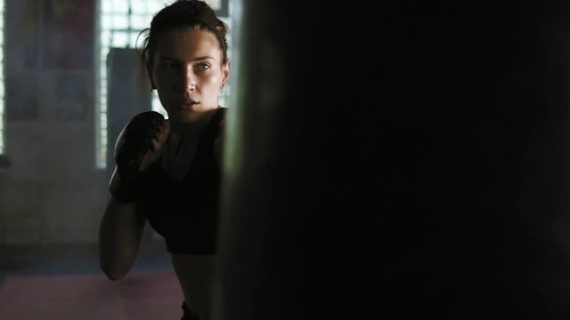 Strong athletic female kickboxer hits a boxing bag with her leg. She is exercising with a boxing bag in dark gym with smoke