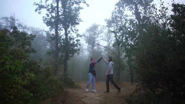 two male friends meeting in foggy wood to play in hacky sack greeting each other giving high five walking along path towards camera in slow motion