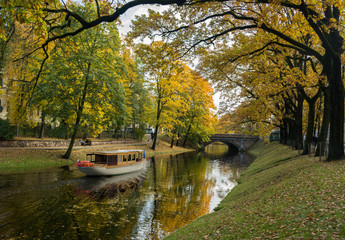 Fototapeta na wymiar Ancient wooden boat on city river canal. Yellow foliage in the autumn park. Riga, Latvia.